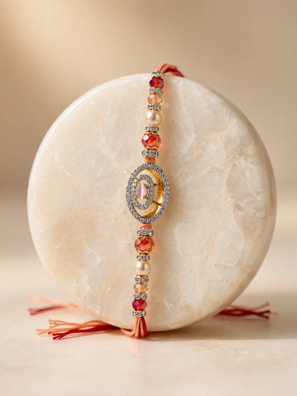 A beaded Rakhi bracelet with red-orange beads, white faux pearls, and a central gold-toned oval charm with clear stones, draped over a round cream stone display on a warm beige background.